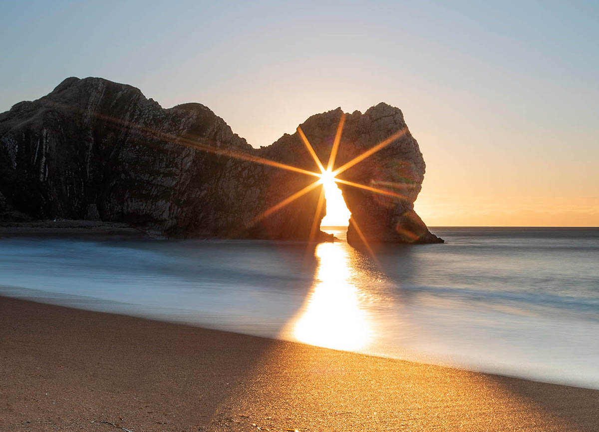 Sunburst through Durdle Door arch at sunrise, Dorset, Jurassic coast, England. Buy a fine art print.